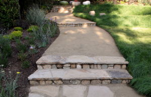 Shallow, deep stairs of flag and cobblestone mirror the earth forms of the IdealMow covered amphitheater, also routing water into the planted areas of the garden and the bioswale. Permeable materials ensure rainwater and irrigation overflow seep back into the soil to nurture the garden.